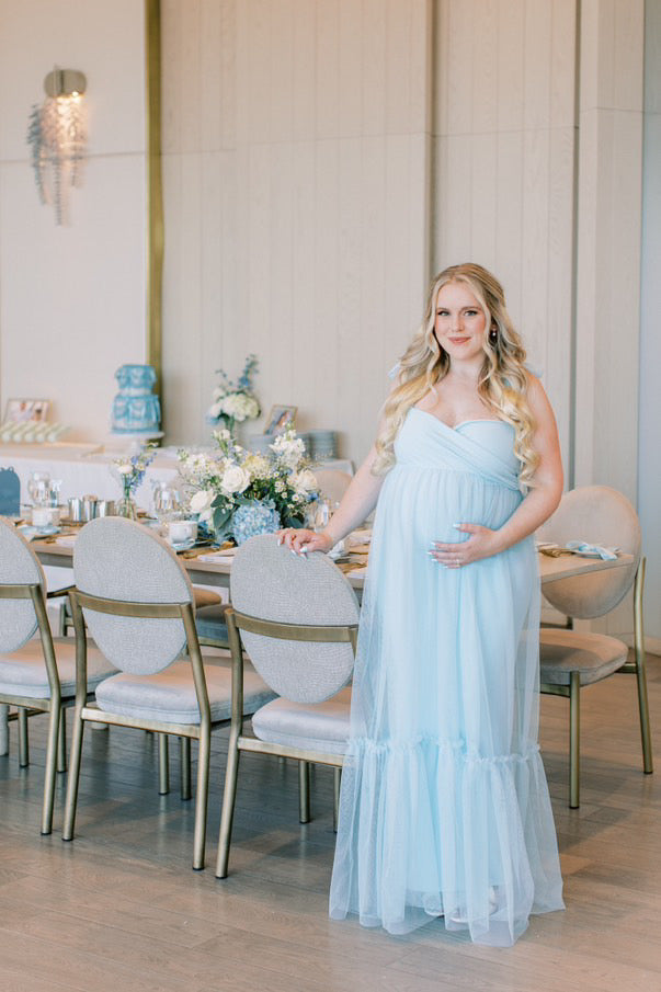 A pregnant woman wearing a blue mesh tulle gown with a sweetheart top design and flowey sheer overlay.